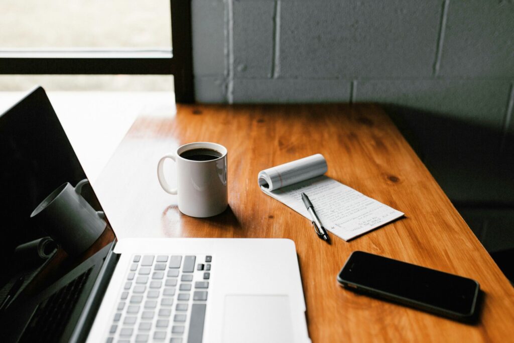 An open silver MacBook laptop sits on a wooden countertop next to a cup of coffee, a notepad, and a smartphone.